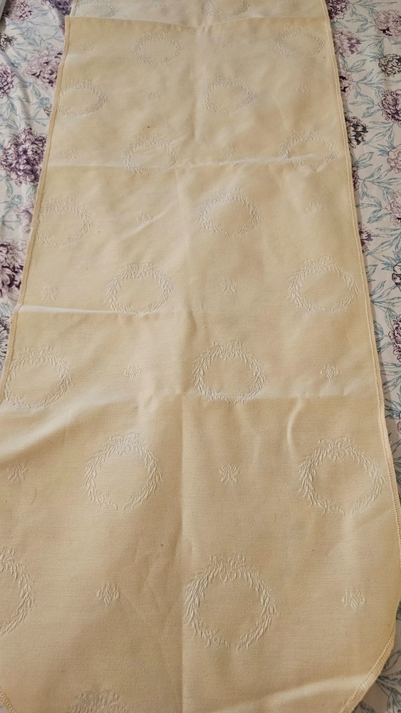 Cream colored table runner with tassled ends and wreath pattern 66 x 13 1/2 in